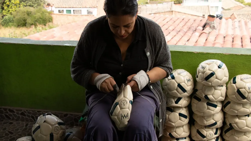 An artisan hand-stitching a Hayworth ball on a balcony in Monguí, finished balls stacked beside her.